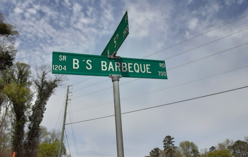 Street sign for B's Barbeque at the intersection of SR 1204 and 5th Street, with trees and a cloudy sky in the background.