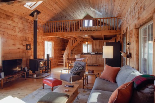 Cozy wooden cabin interior with a living area, kitchen, and lofted bedroom, featuring a fireplace and comfortable seating.