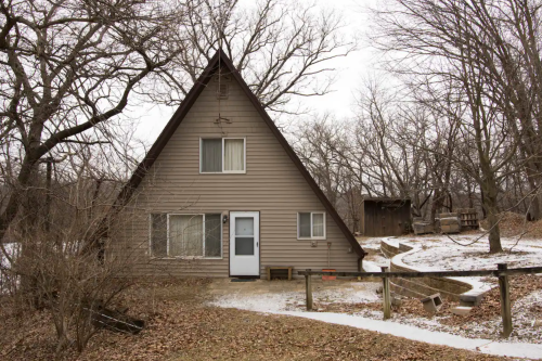 A triangular-shaped house surrounded by bare trees and a snowy path, with a small shed in the background.