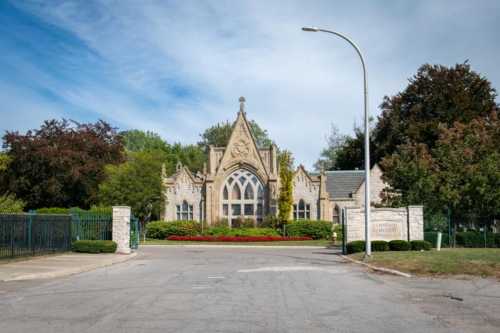 A stone building with gothic architecture, surrounded by trees and colorful flowers, at the entrance of a cemetery.