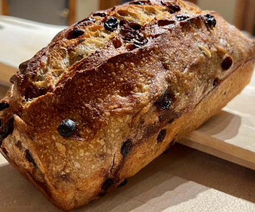 A freshly baked loaf of bread with a golden crust and dark raisins on a wooden surface.