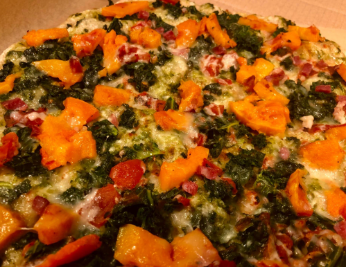 A close-up of a pizza topped with kale, butternut squash, and bacon on a golden crust.
