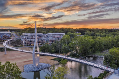 A modern pedestrian bridge spans a river, surrounded by trees, with a vibrant sunset in the background.