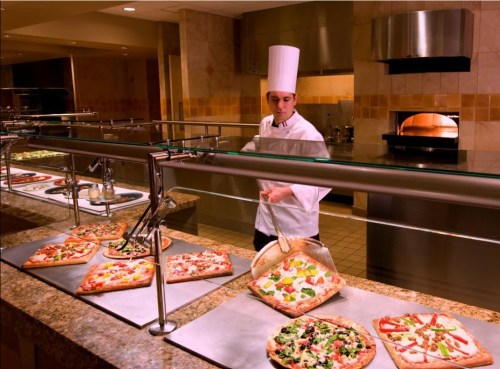 A chef in a white uniform serves pizza from a buffet in a restaurant kitchen. Various pizzas are displayed on the counter.