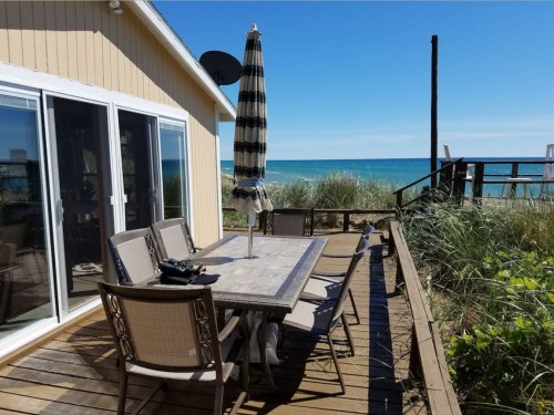 A beachside patio with a dining table and chairs, overlooking the ocean and sandy shore under a clear blue sky.