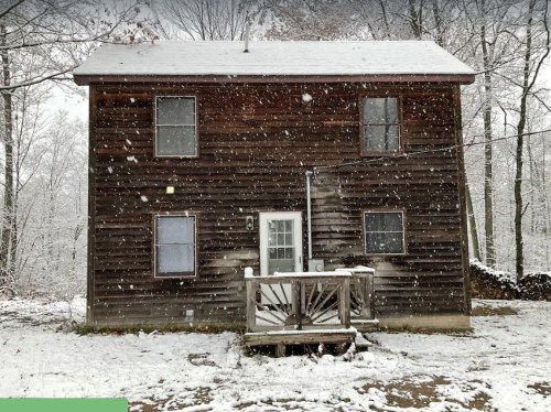 A wooden house with snow on the ground and falling snowflakes, surrounded by trees in a winter landscape.