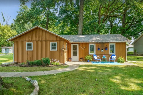 A cozy wooden house surrounded by greenery, featuring a front porch with blue chairs and a well-maintained lawn.