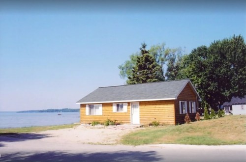A small wooden cabin by a lake, surrounded by trees and grass, under a clear blue sky.