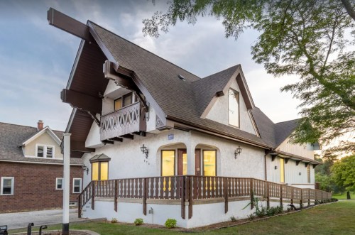 A charming two-story house with a steep roof, balcony, and wooden railing, surrounded by greenery at sunset.