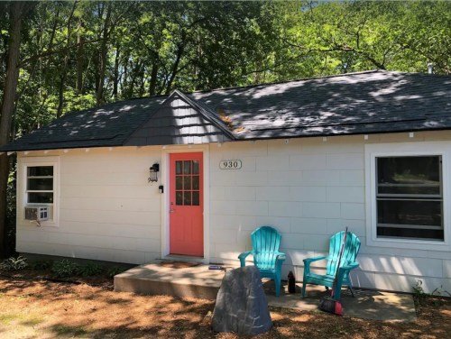 A small white house with a pink door, two blue chairs on the porch, and surrounded by trees.