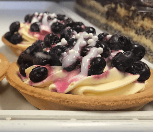 Two tartlets filled with creamy yellow frosting, topped with blackberries and white sprinkles, displayed in a pastry case.
