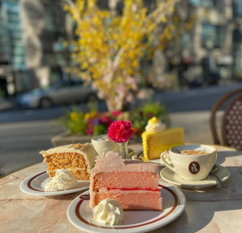 A table with three colorful cakes and a cup of coffee, surrounded by flowers and a sunny outdoor setting.