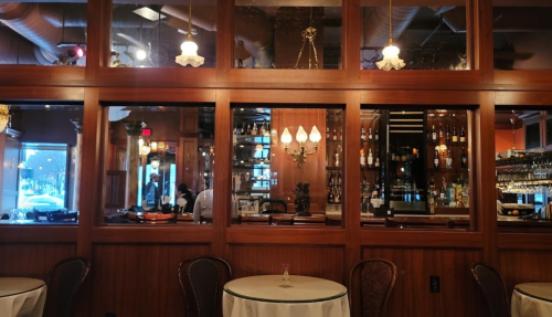 Interior of a restaurant featuring wooden paneling, a bar with bottles, and elegant lighting. Tables are set for dining.