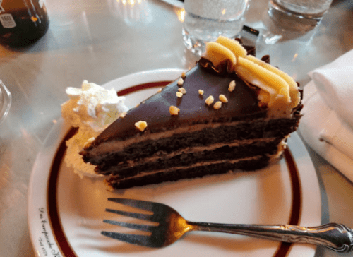 A slice of chocolate cake with layers, topped with chocolate ganache and banana slices, served with whipped cream.