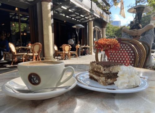 A cup of coffee beside a plate of tiramisu topped with whipped cream, set at an outdoor café.