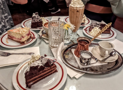 A table with various desserts, including chocolate cake and cheesecake, alongside drinks and a decorative glass.