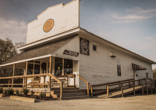 A rustic white general store with a porch, sign, and bakery, set against a clear sky.