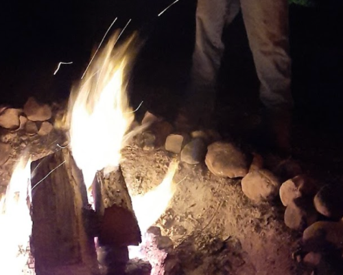 A campfire burns brightly at night, surrounded by rocks, with a person standing nearby.