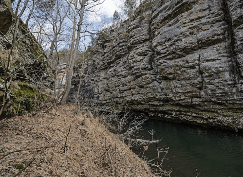 Rocky cliffs surround a calm, greenish water body, with sparse trees and dry grass along the bank.