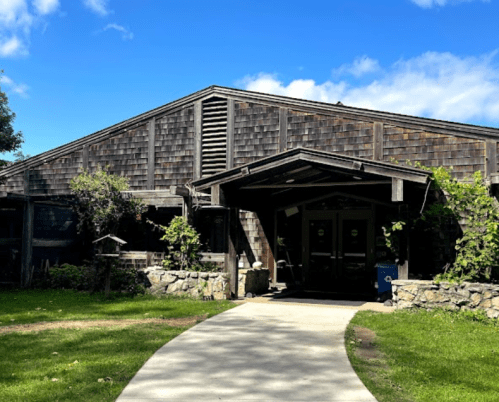 A rustic wooden building with a sloped roof, surrounded by greenery and a paved walkway leading to the entrance.