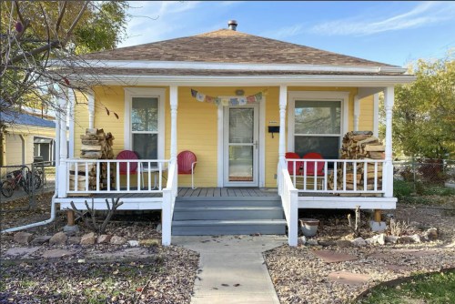 A charming yellow house with a front porch, two red chairs, and stacked firewood on either side, surrounded by a garden.