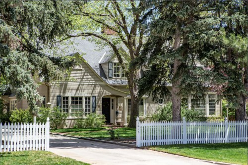 A charming house with a gray roof, surrounded by trees and a white picket fence on a sunny day.
