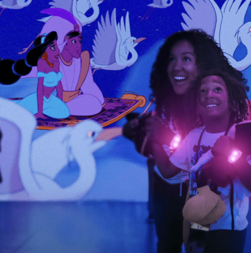 A child and an adult smile while watching a scene from a Disney movie featuring characters on a magic carpet with swans.