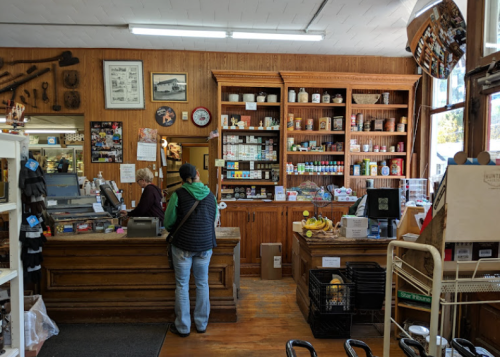 A cozy store interior with wooden shelves filled with jars and products, a cashier, and a customer at the counter.