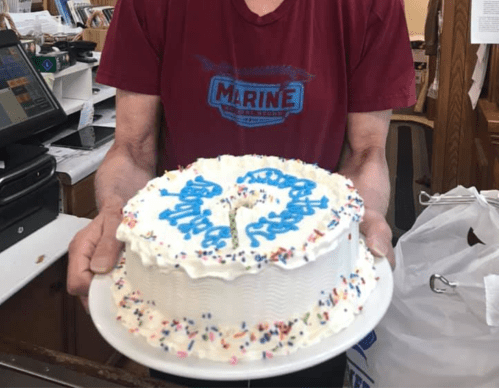 A person holds a decorated birthday cake with blue icing and colorful sprinkles, featuring the number "0" in the center.