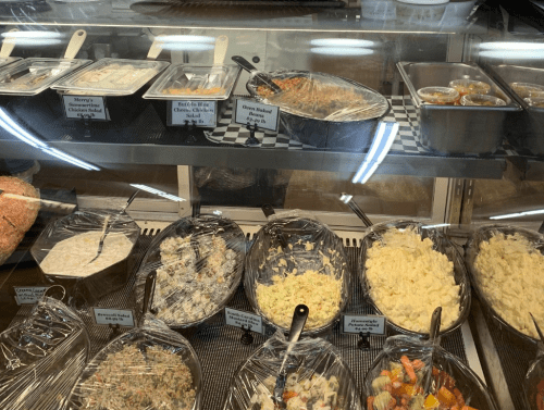 A display case filled with various prepared dishes, including salads, baked items, and sides, all covered with plastic wrap.