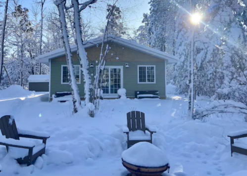 A cozy green cabin surrounded by snow, with chairs and a fire pit in the foreground under a bright winter sun.