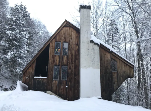 A wooden cabin with a sloped roof, partially covered in snow, surrounded by trees in a winter landscape.