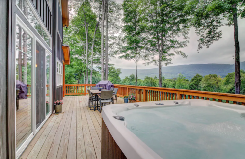 A wooden deck with a hot tub overlooks a scenic view of trees and mountains under a cloudy sky.