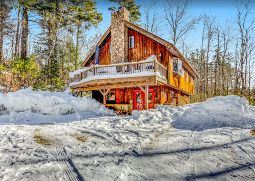 A cozy wooden cabin surrounded by snow, featuring a stone chimney and a front porch, set in a winter landscape.