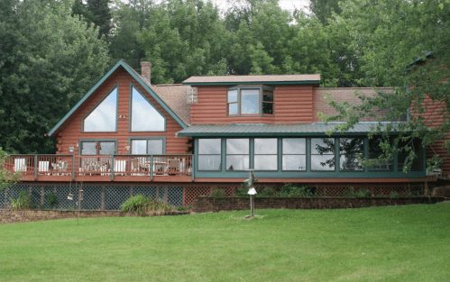 A large wooden house with a deck, surrounded by greenery and trees, set on a grassy lawn.