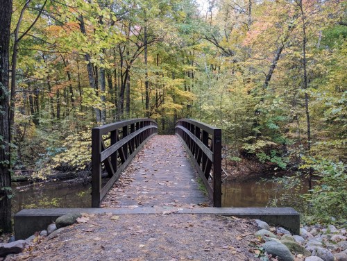 A wooden bridge spans a calm stream, surrounded by vibrant autumn foliage and trees.