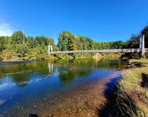 A wooden bridge spans a calm river, surrounded by lush green trees under a clear blue sky.