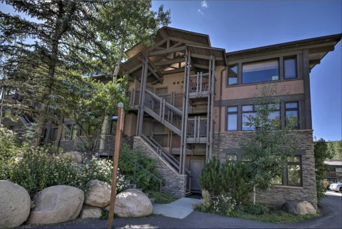 A modern building with wooden accents, surrounded by trees and large rocks, featuring a staircase and multiple windows.