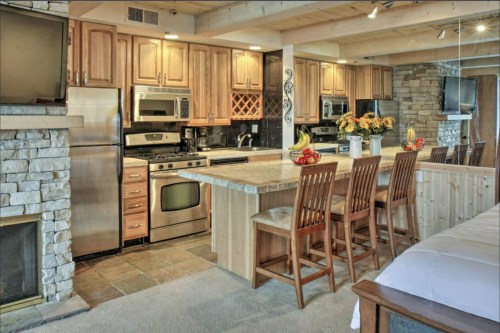 Modern kitchen with wooden cabinets, stainless steel appliances, a stone fireplace, and a large island with bar stools.