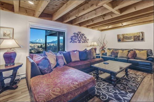 Cozy living room with a sectional sofa, coffee table, and large windows showcasing a mountain view. Warm lighting and decor.