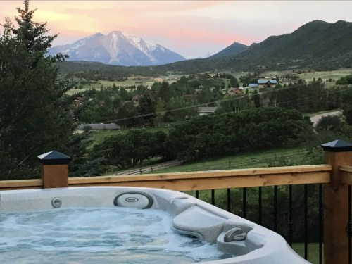 A hot tub on a deck overlooks a scenic mountain landscape at sunset, with lush greenery and a small town below.
