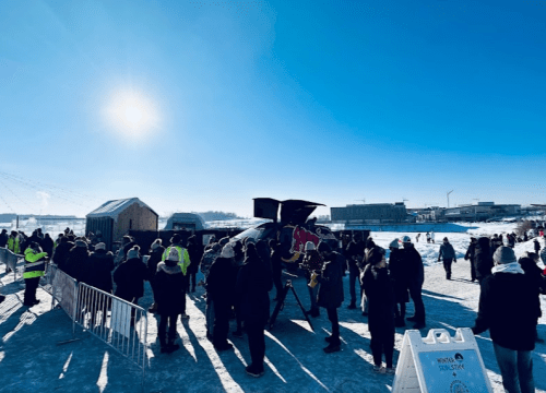A crowd gathers outdoors on a sunny winter day, with snow and ice surrounding them. A stage is set up in the background.