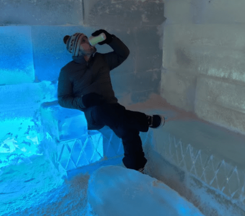 A person sits on an ice bench, drinking from a bottle in a brightly lit ice room.