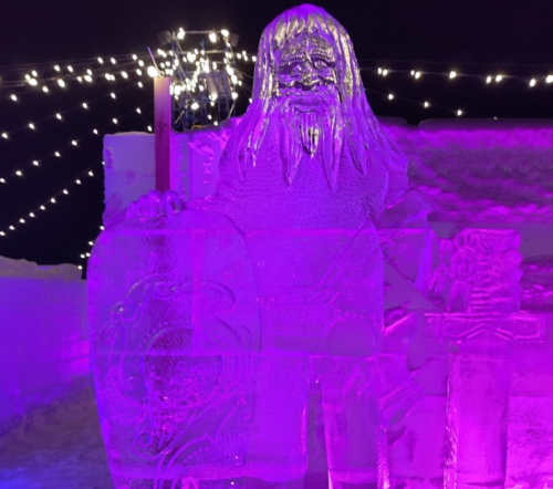 An illuminated ice sculpture of a bearded figure holding a candle, surrounded by festive lights.