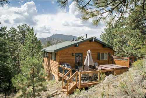 A wooden cabin on a hillside surrounded by trees, with a deck, hot tub, and mountain views under a cloudy sky.