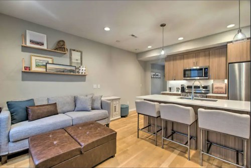 Modern living space featuring a gray sofa, ottoman, and a kitchen with bar stools, sleek cabinets, and stainless appliances.