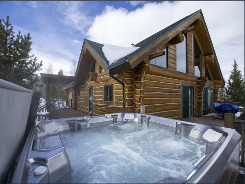 A log cabin with a hot tub in the foreground, surrounded by snow and trees under a blue sky.