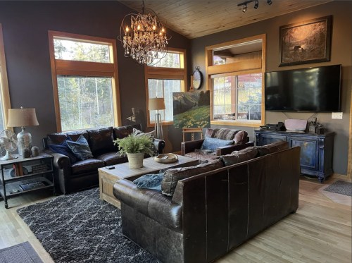 Cozy living room with large windows, a chandelier, leather sofas, a wooden coffee table, and a TV.