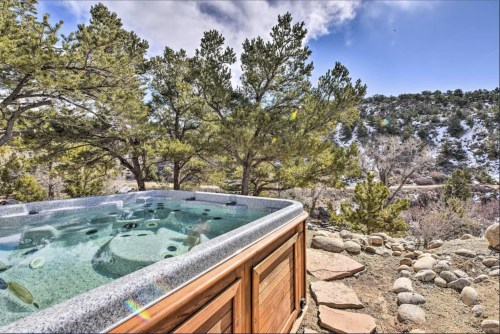 A hot tub surrounded by trees and rocky terrain, with a scenic view of mountains and a blue sky.