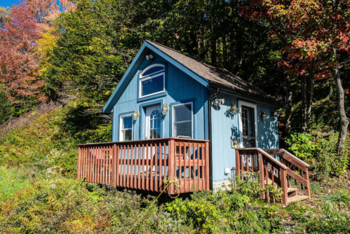 A small blue cabin with a wooden deck, surrounded by colorful autumn foliage.